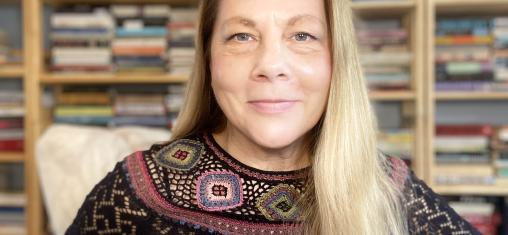 A woman with blond hair and a black patterned sweater sits in front of bookshelves.