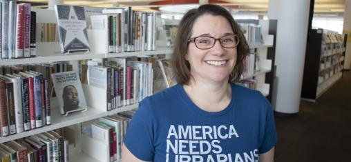 A woman in a blue t-shirt that reads "America Needs Librarians" smiles in front of book shelves.