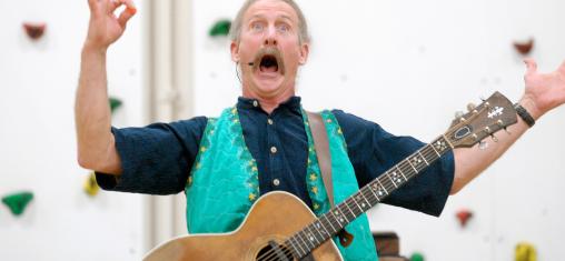 A man in a blue vest, wearing a guitar, makes an open-mouthed expression while raising his arms.