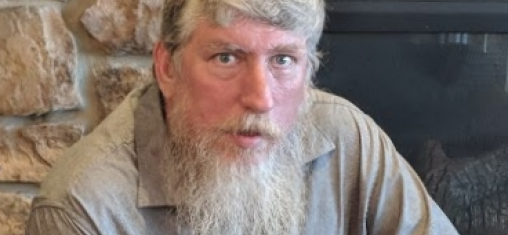 Joseph Engler has a long beard and white hair and sits in front of a fireplace.