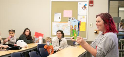 A woman holds up a children's book she is reading to a room of parents and children.