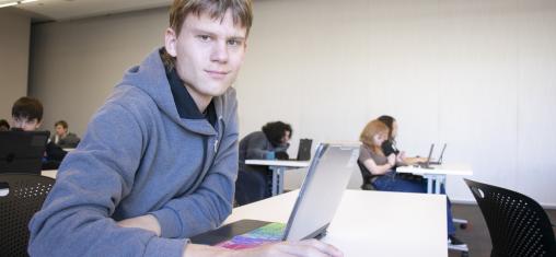 A teen boy looks at the camera while working on a laptop.