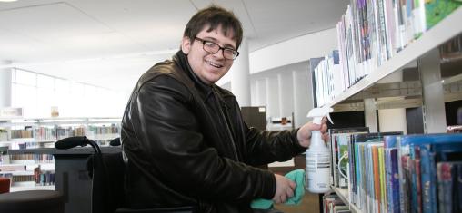 A man in a wheelchair smiles at the camera while holding cleaning supplies in front of bookshelves.