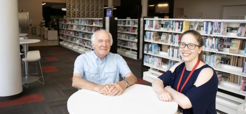 An older man and a woman smile at the camera in front of book shelves in the library.