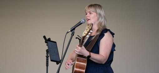 A woman holding a guitar sings into a microphone.