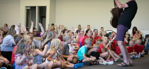 A man holds a guitar and sings in front of a crowd of seated children.