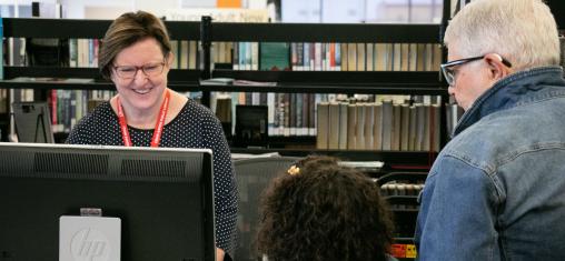A woman and girl look at a library worker who is helping them from behind a computer.