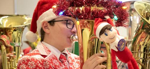 A young man wearing a Santa hat and Christmas sweater plays a tuba that is decorated with garland.