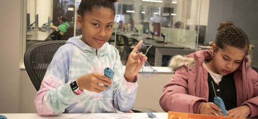A girl smiles as she and another girl use Doodle Pens at the library.