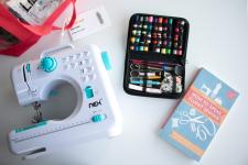 A mini sewing machine and kit with thread and sewing tools sit next to a book on a white table.