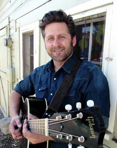 Musician Chad Elliott holding a guitar against the background of a white house