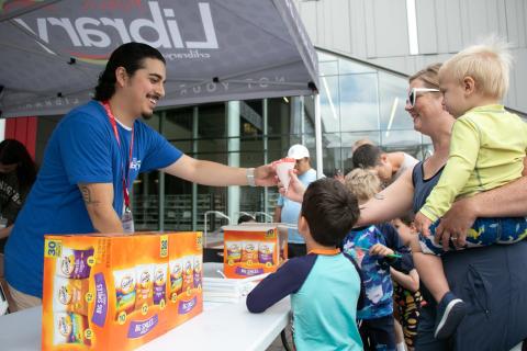 A man hands a woman and children a snow cone in front of the Downtown Library.