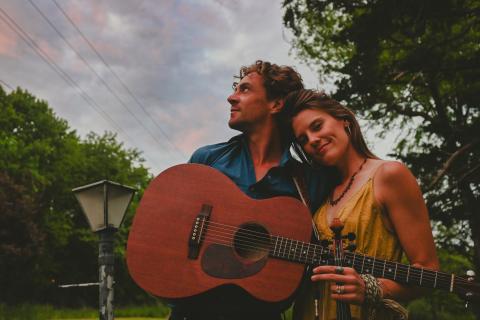 Musicians Forrest and Margaret McCurren standing together against a blue sky. Forrest is holding a guitar, Margaret is holding a fiddle.