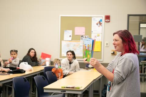 A woman reads a picture book to a room of children and parents.