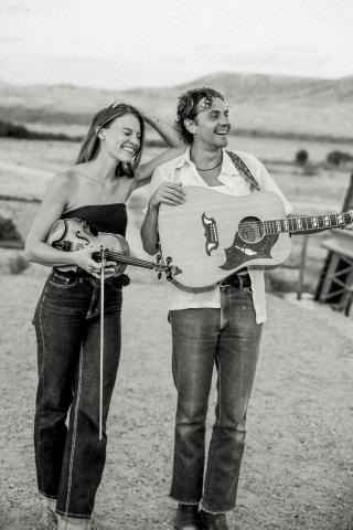Woman holding a fiddle leaning on man holding a guitar set against the backdrop of open plains with mountains in the background