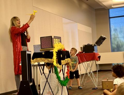 Mikayla Oz, wearing a long red coat, holds up a balloon dog while a boy laughs.