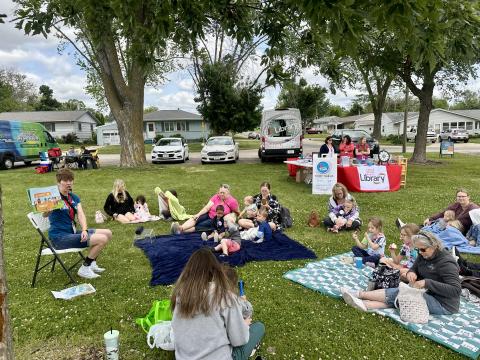 A librarian reads a book to children and adults sitting on blankets and the grass at a park, with a library activity table in the background.
