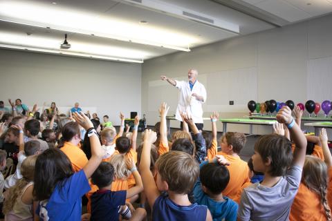 A man in a white lab coat calls on children who are raising their hands.