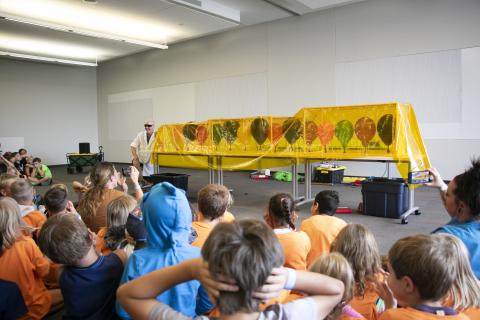 Children cover their ears as a man in a white lab coat prepares an experiment that will pop a line of balloons.