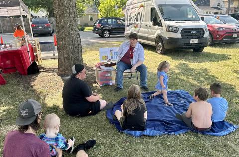 A man reads a book to watching children sitting with some adults on a blue blanket in Cleveland Park.