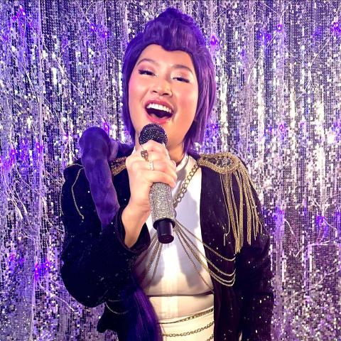 A woman with a purple wig and purple jacket with gold epaulettes sings into a bejeweled microphone in front of a sequined backdrop.
