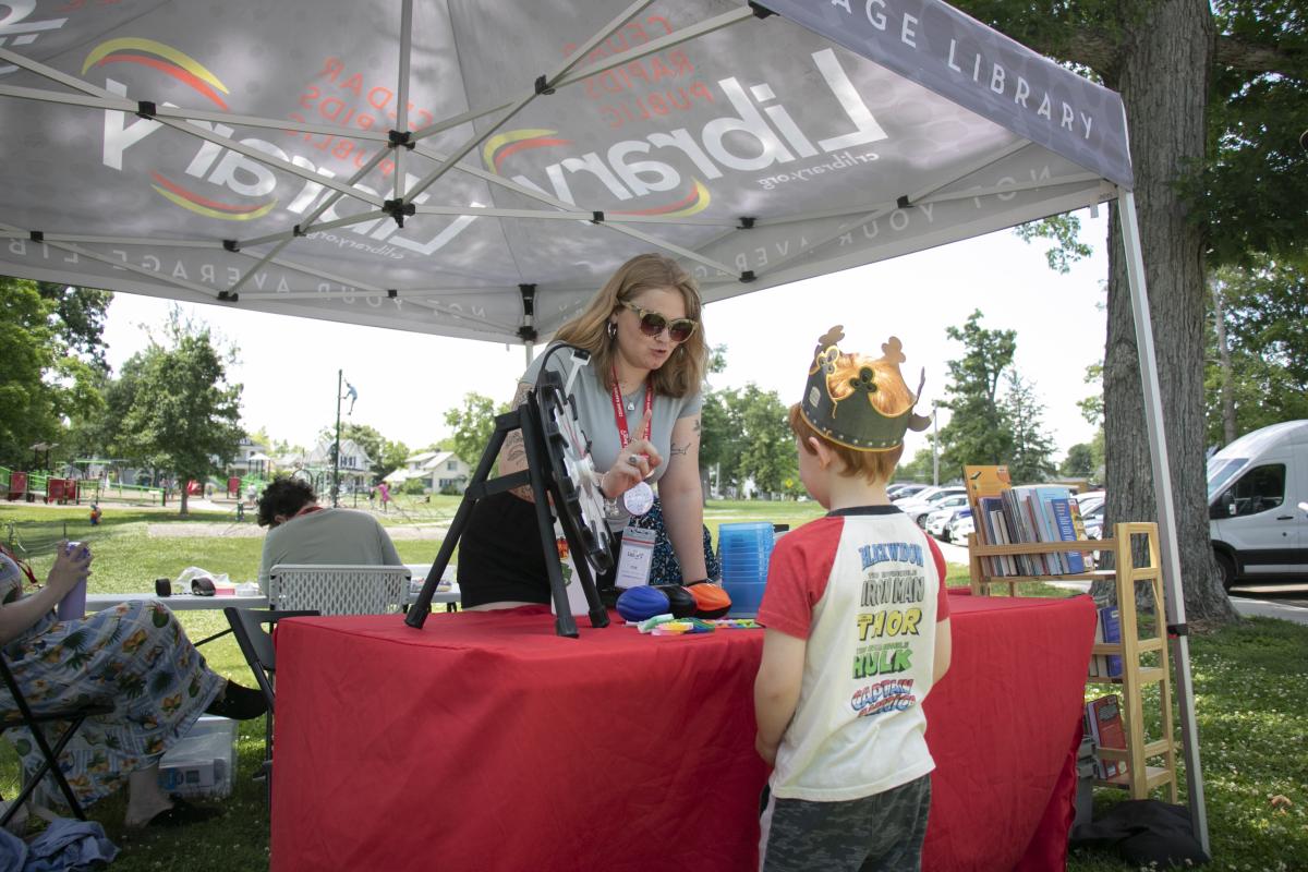 A boy in a crown talks to a woman at a table in a Cedar Rapids Public Library tent in Bever Park.