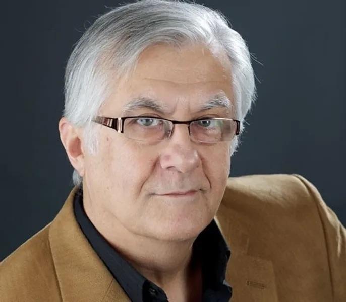 An older man with white hair and glasses, wearing a tan sport coat, looks at the camera.