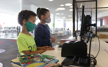 Two children with masks checking out stack of books