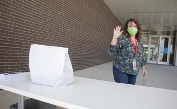A white paper bag sits on a table while a woman in a mask waves.