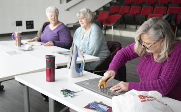 A woman in a pink sweater cuts fabric on a table.