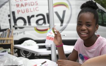 A girl smiles while holding a craft in front of the Mobile Technology Lab.
