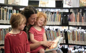 A woman reads to a girl on her lap while another girl looks on.